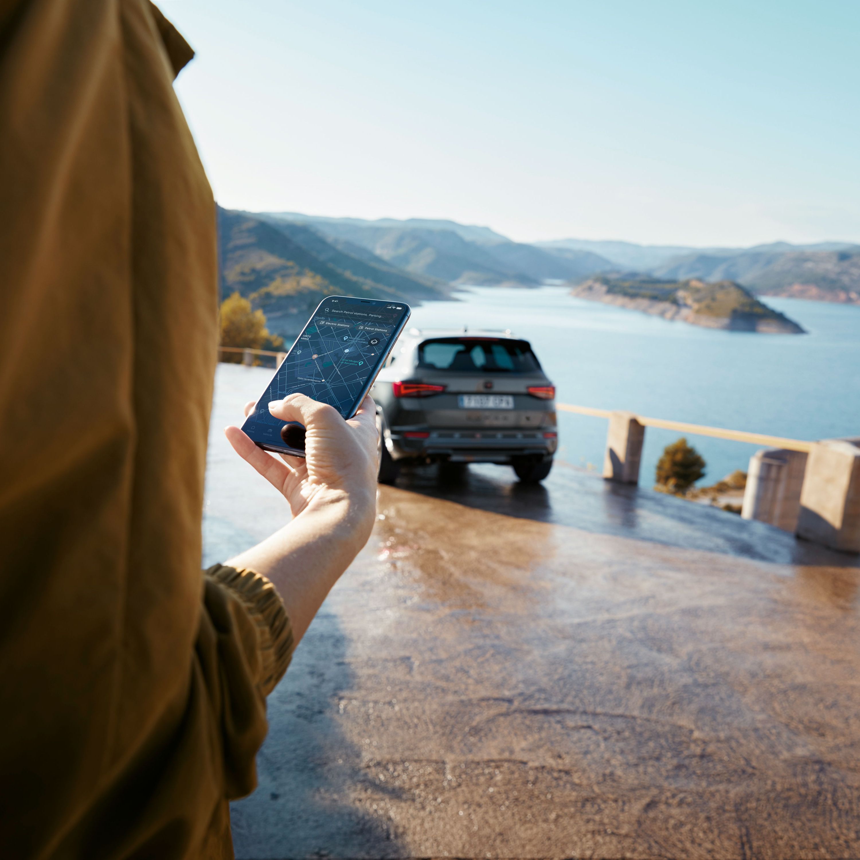 Eine Person hält ein Smartphone mit dem CUPRA Ateca im Hintergrund, der in der Nähe einer malerischen Aussicht auf Berge und Wasser parkt, was eine vernetzte Welt symbolisiert.