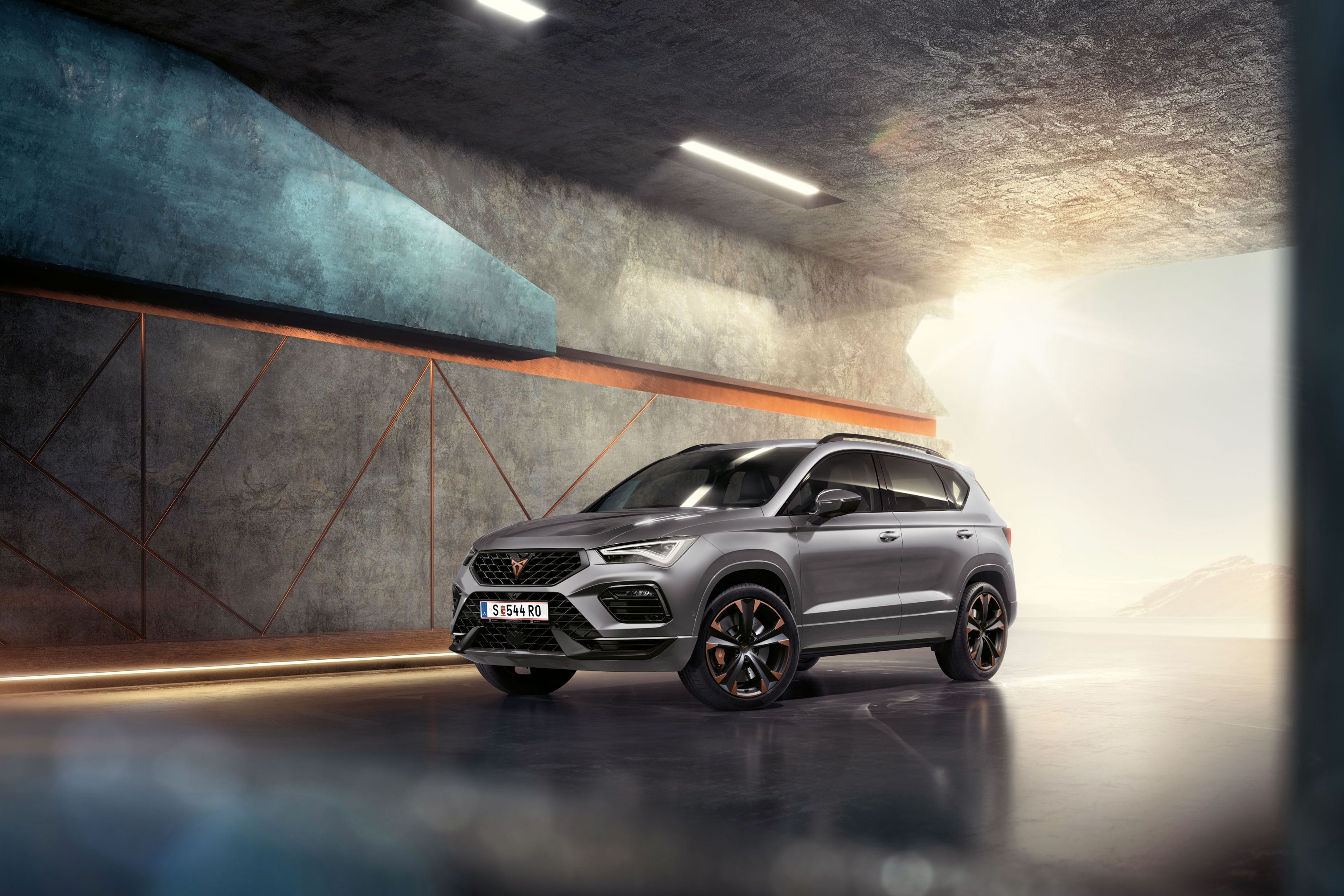 Ein 2024 Cupra Ateca in Graphitgrau, ein sportlicher und kompakter Geländewagen, der in einem modernen, industriellen Umfeld geparkt ist. Das Fahrzeugdesign zeichnet sich durch aerodynamische Linien, LED-Scheinwerfer und markante Leichtmetallfelgen mit Kupferakzenten aus. Der Hintergrund besteht aus einem geometrischen Wanddesign, das mit dem eleganten Fahrzeugdesign kontrastiert.