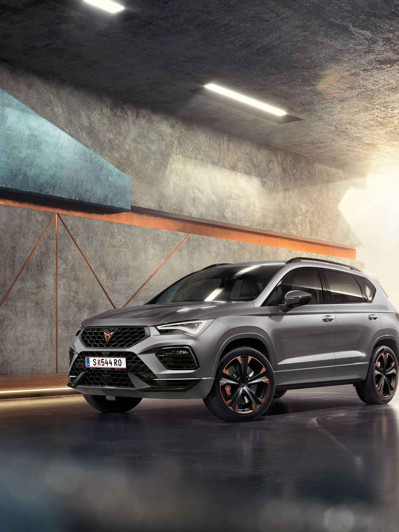 Ein 2024 Cupra Ateca in Graphitgrau, ein sportlicher und kompakter Geländewagen, der in einem modernen, industriellen Umfeld geparkt ist. Das Fahrzeugdesign zeichnet sich durch aerodynamische Linien, LED-Scheinwerfer und markante Leichtmetallfelgen mit Kupferakzenten aus. Der Hintergrund besteht aus einem geometrischen Wanddesign, das mit dem eleganten Fahrzeugdesign kontrastiert.