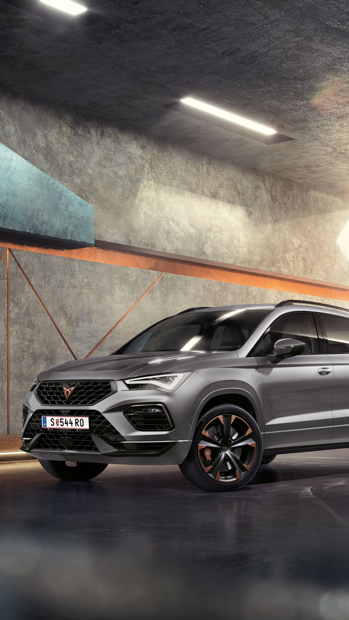 Ein 2024 Cupra Ateca in Graphitgrau, ein sportlicher und kompakter Geländewagen, der in einem modernen, industriellen Umfeld geparkt ist. Das Fahrzeugdesign zeichnet sich durch aerodynamische Linien, LED-Scheinwerfer und markante Leichtmetallfelgen mit Kupferakzenten aus. Der Hintergrund besteht aus einem geometrischen Wanddesign, das mit dem eleganten Fahrzeugdesign kontrastiert.
